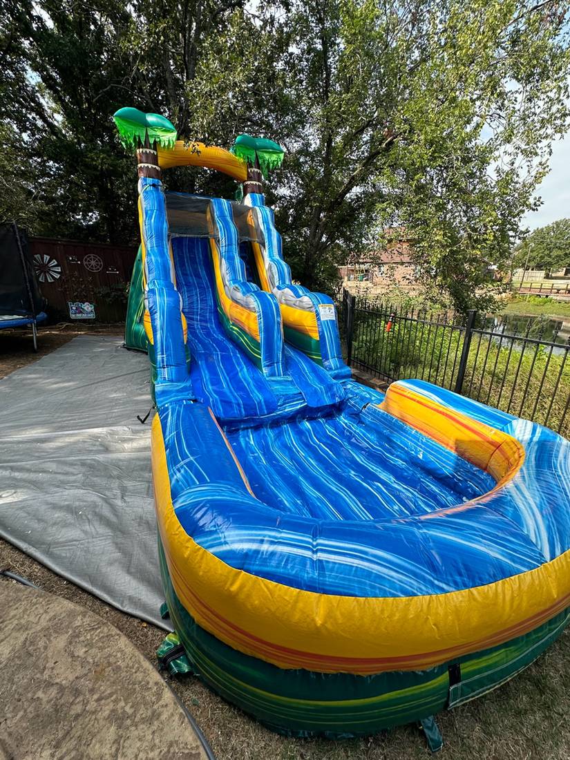 Tall blue and green double lane water slide with palm tree tops and a splash pool set up in a yard with trees behind