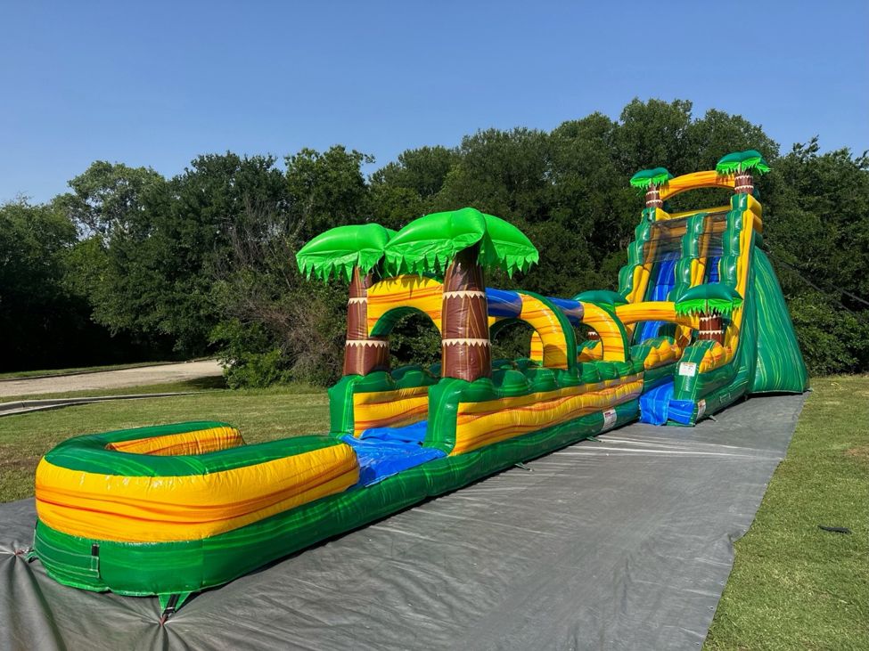 Long green and yellow tropical palm tree water slide with a splash pool set up on a lawn with trees in the background