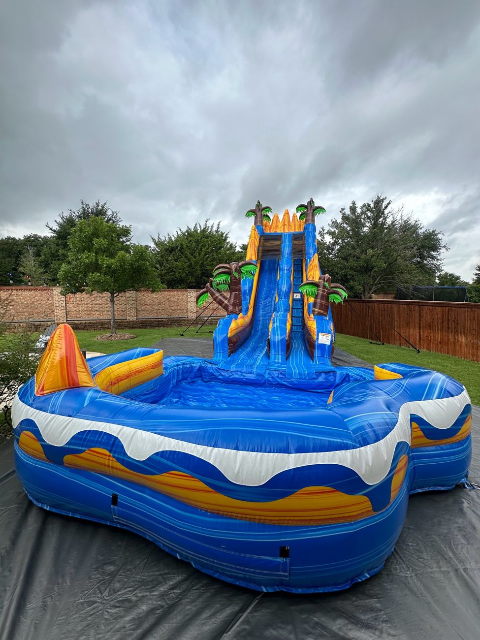 Blue tropical water slide with palm tree tops and a splash pool set up in a backyard