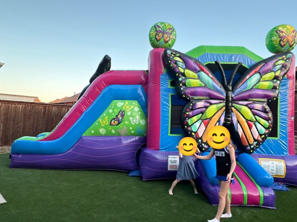 Purple, pink and blue butterfly-themed combo bounce house with a slide on artificial turf in a backyard, with two kids playing in front (faces covered with smiley stickers)