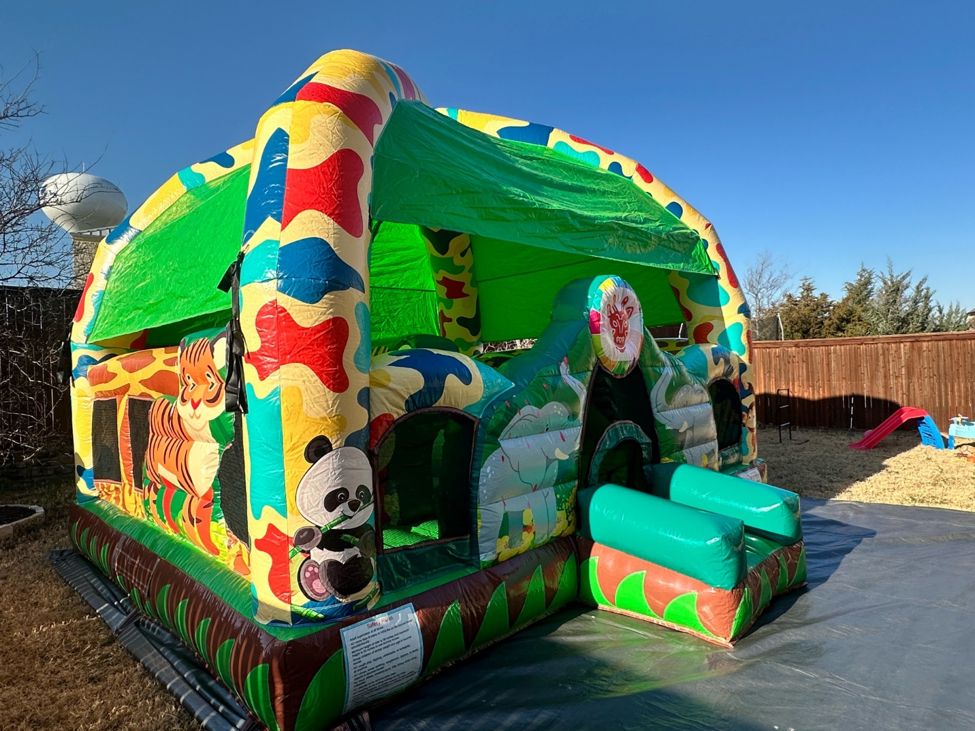 Colorful jungle animal bounce house with a small slide set up on a black tarp in a backyard