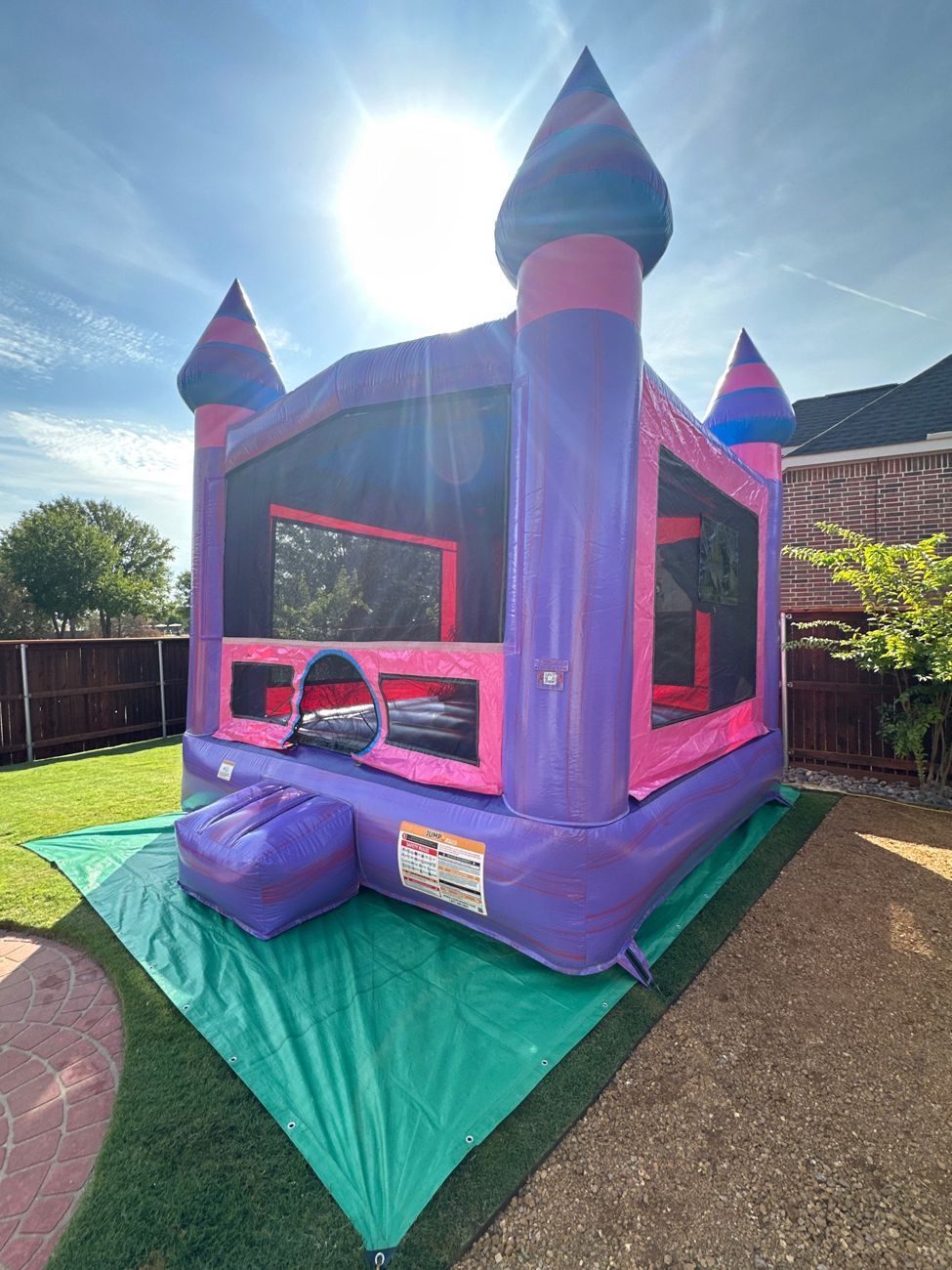 Purple and pink castle bounce house set up on a green tarp in a backyard with a wood fence