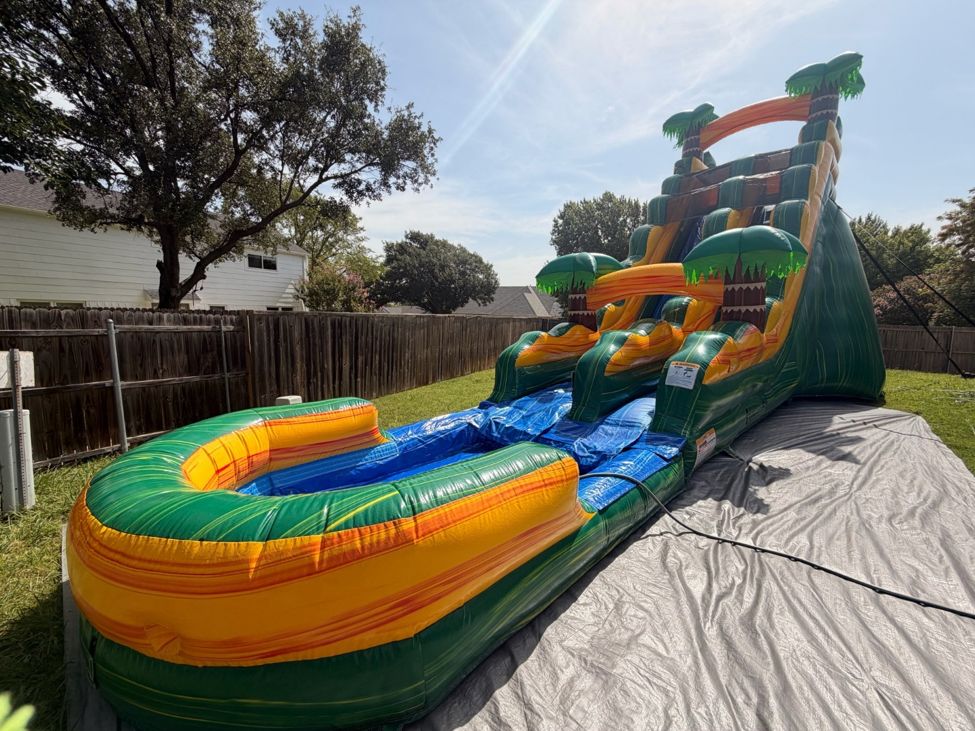 Tall green and orange tropical water slide with a splash pool set up on a lawn beside a wood fence