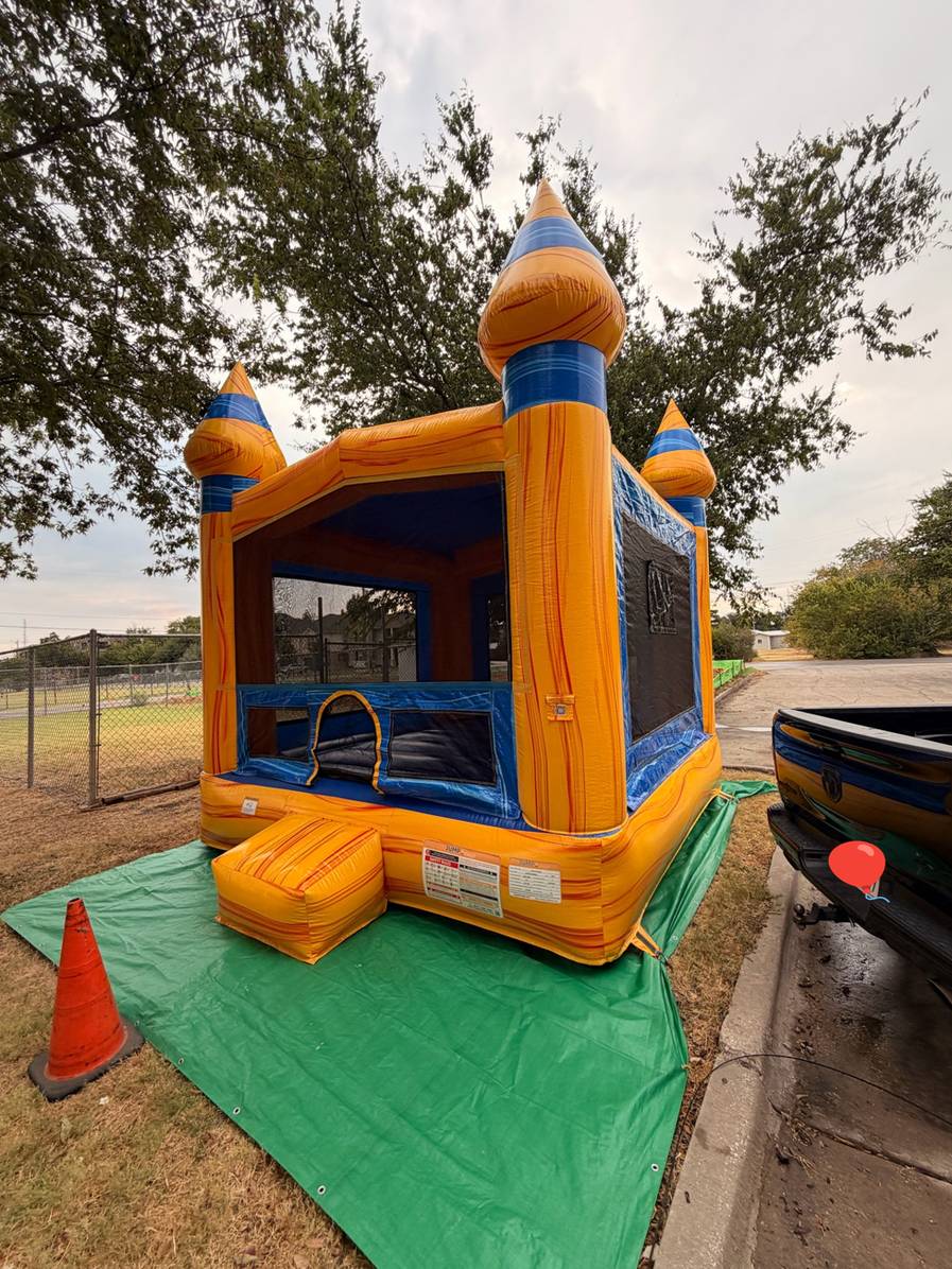 Orange and blue castle bounce house set up on a green tarp on a grass strip next to a paved parking area