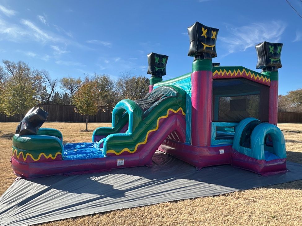 Teal and pink bounce house combo with a curved slide set up on a gray tarp on a dry grass lawn