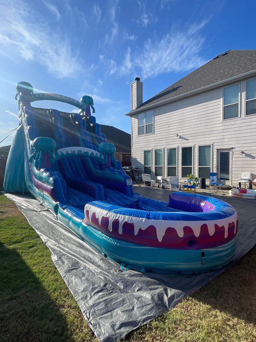 Tall blue and teal water slide with a splash pool set up on a gray tarp in a backyard beside a house