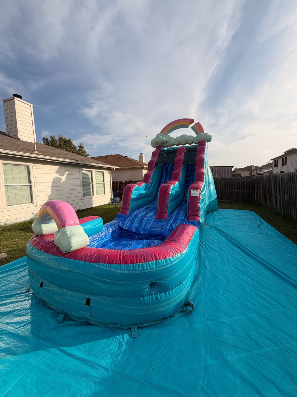 Pink and teal water slide with a rainbow topper set up on a teal tarp in a backyard