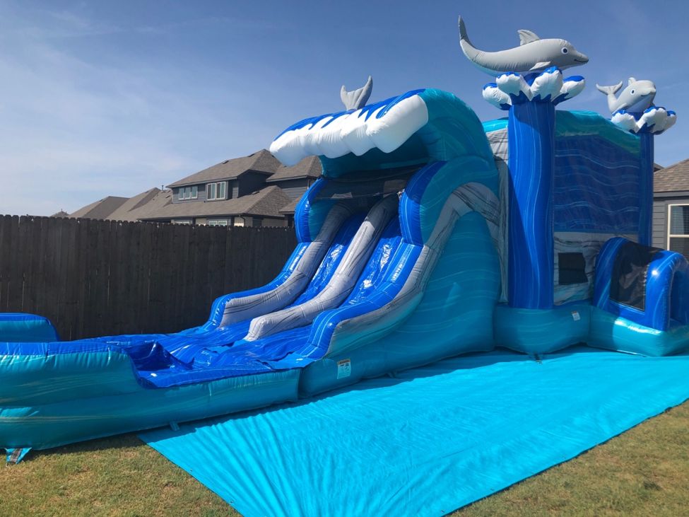 Blue and teal wave-themed water slide combo with two slide lanes set up on a lawn beside a wood fence