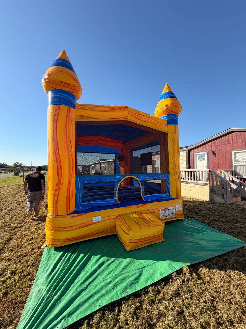 Orange and blue castle bounce house set up on a green tarp on a grassy lot, with a person walking away in the background