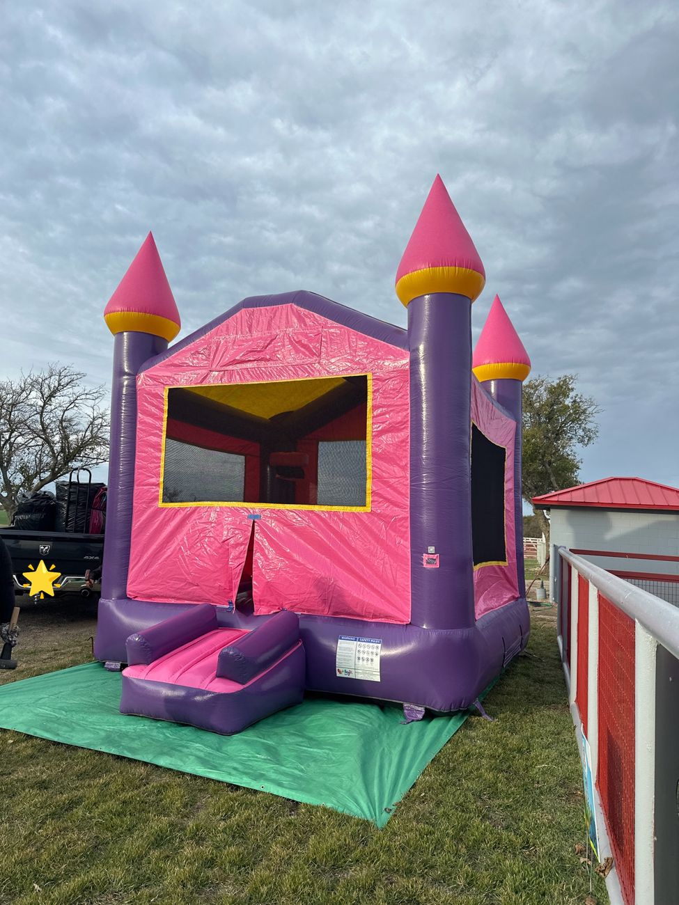 Pink and purple castle bounce house set up on a green tarp on grass under a cloudy sky