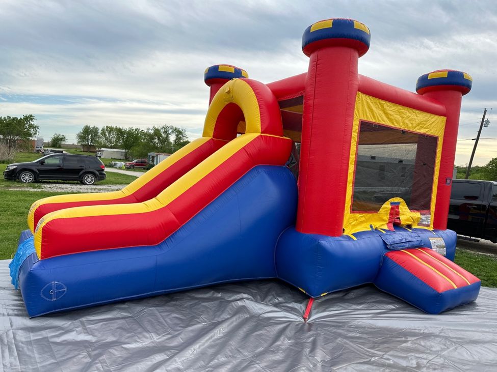 Red, yellow and blue bounce house with a wide slide set up on a gray tarp on a lawn