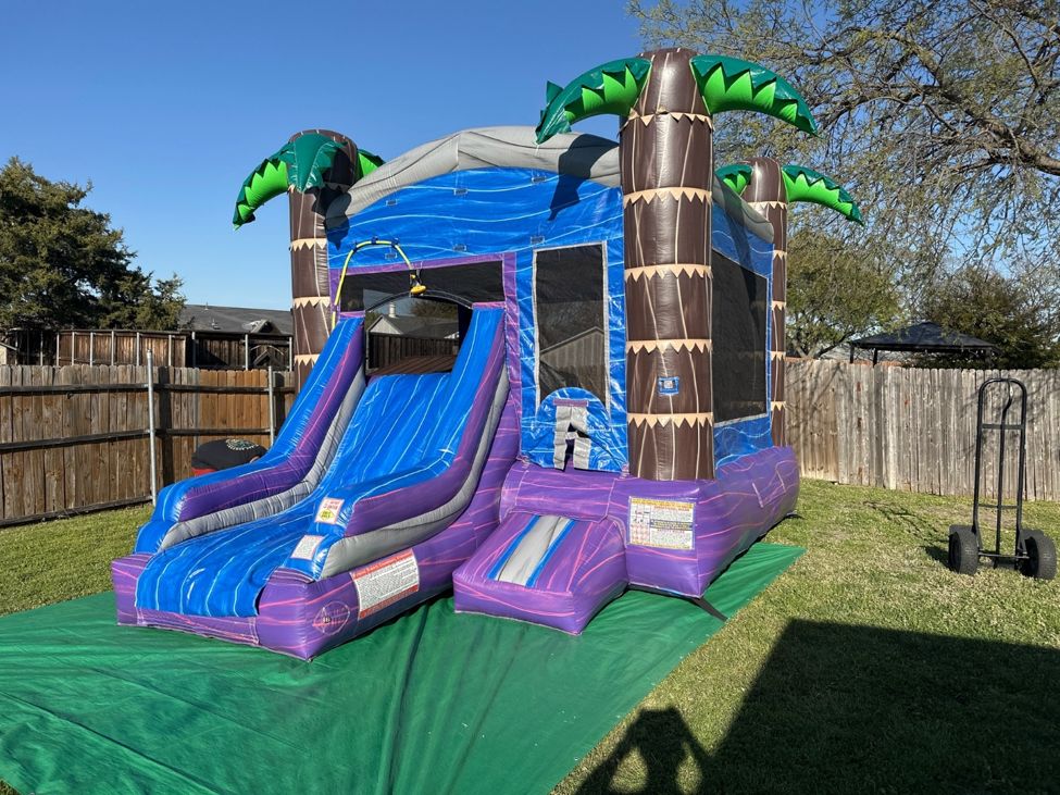 Purple and blue palm tree themed combo bounce house with a slide set up on a green tarp in a fenced backyard